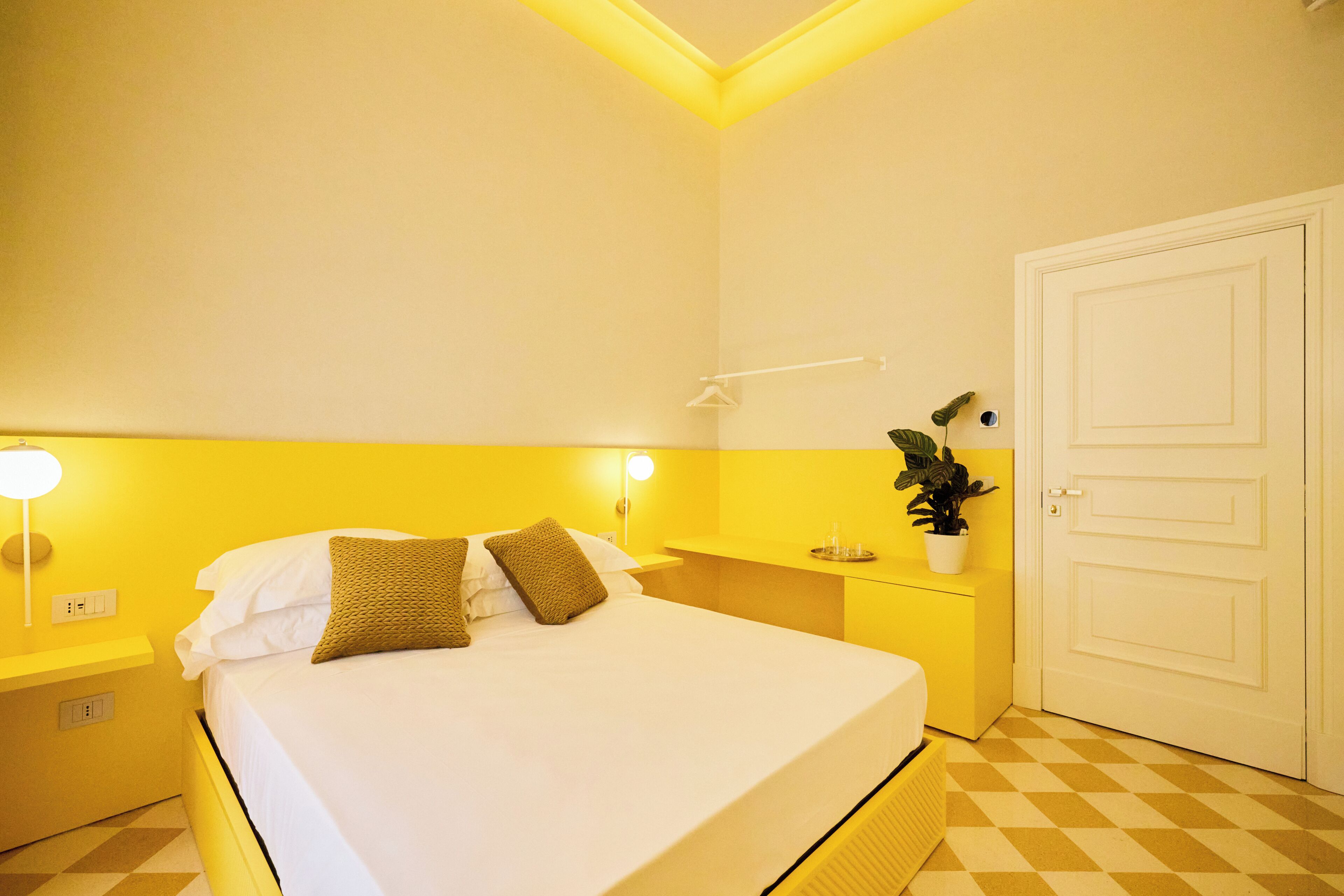 A hotel room with a bed, bedside tables, a nightstand, a wall-mounted lamp, a potted plant, and a door.
