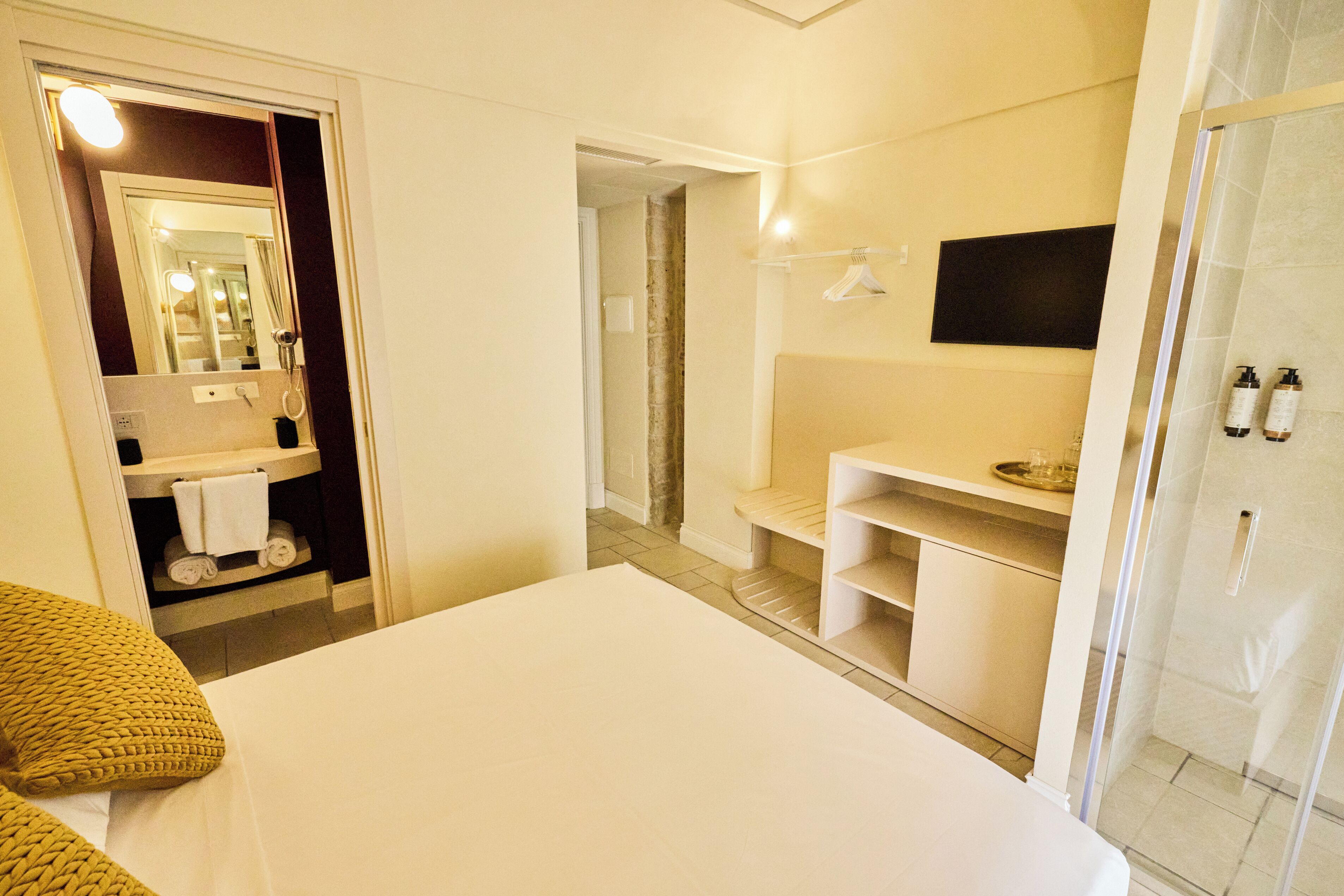 A hotel room with a bed, a bathroom with a shower, a mirror, and a shelf with toiletries.
