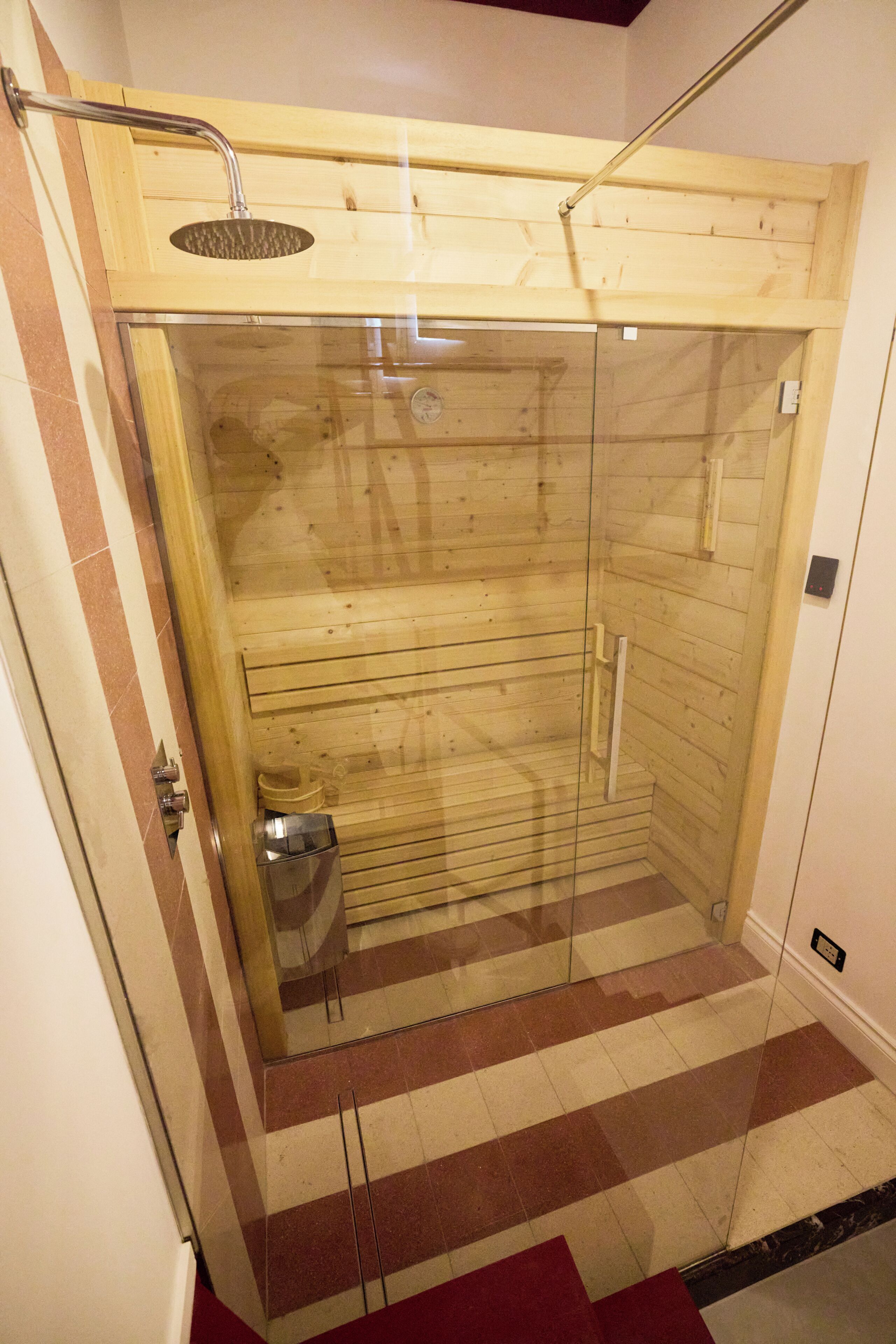 A glass-enclosed wooden sauna with a showerhead and a small heater.