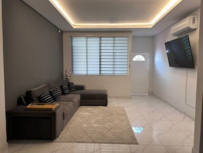 A modern living room with a sectional sofa, a flat-screen TV mounted on the wall, and a large window with blinds.