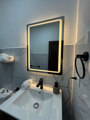 Un baño con un lavamanos blanco, un espejo con luz, una toalla en la pared y un grifo negro.
