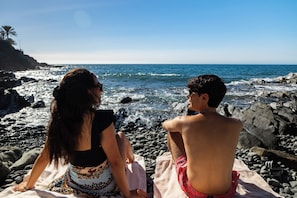 Two people sitting on a rocky beach with the ocean in the background.