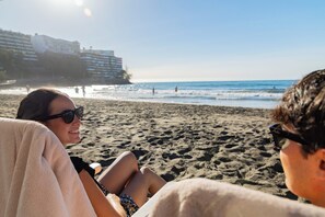 A beach scene with people relaxing on the sand, a clear sky, and buildings in the background. - Sun & Figs (San Bartolomé de Tirajana)