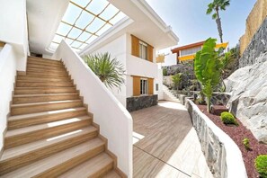 A modern house with a staircase leading to a second floor, a patio area with a stone wall, and a view of a neighboring building. - Sun & Figs (San Bartolomé de Tirajana)