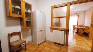 Eine kleine Küche mit Kühlschrank, Holzschränken, einem Stuhl und einem Fenster.