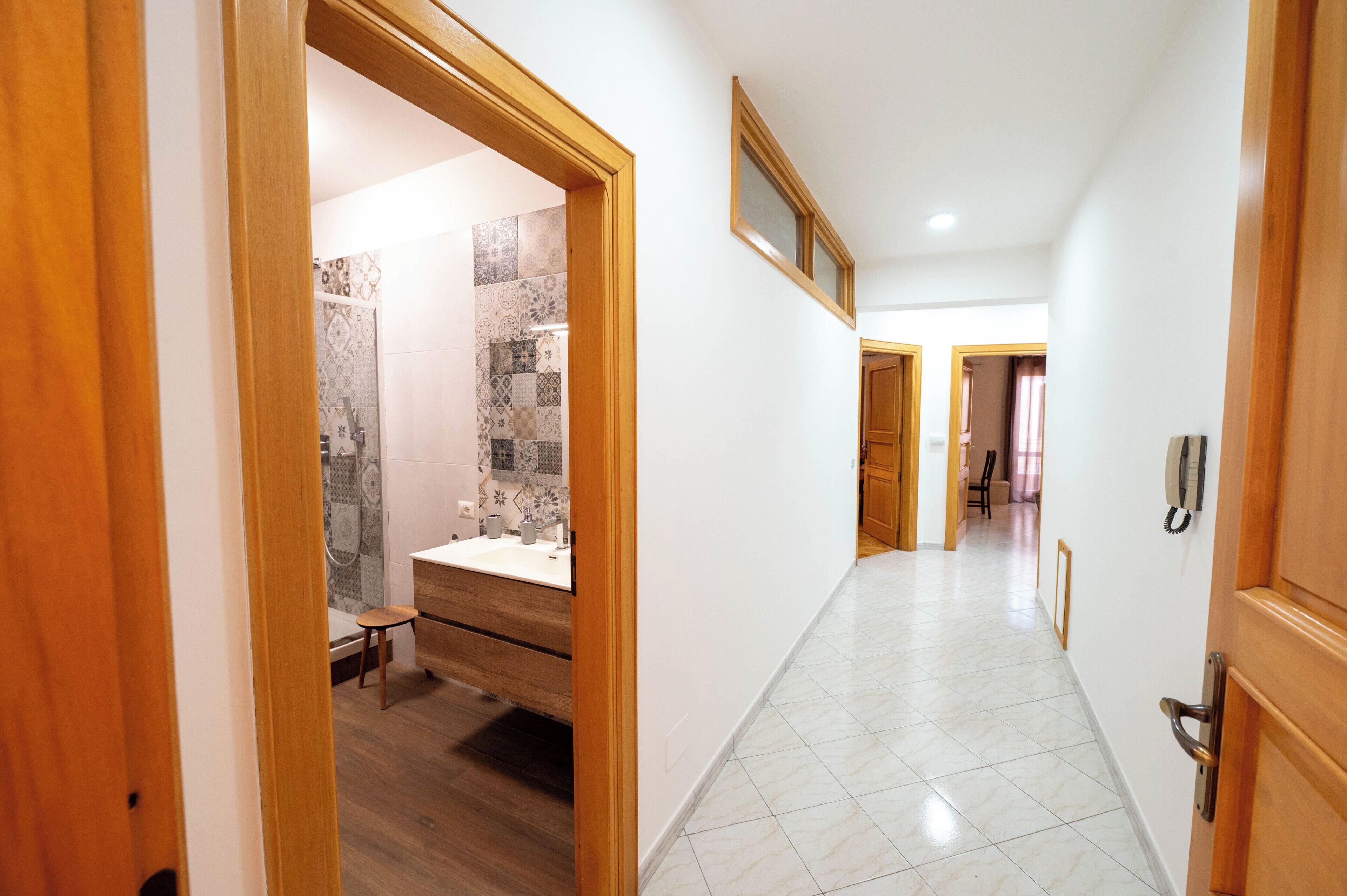 Un couloir d’hôtel avec un cadre de porte en bois, une salle de bain avec une douche et un lavabo, et un sol carrelé.