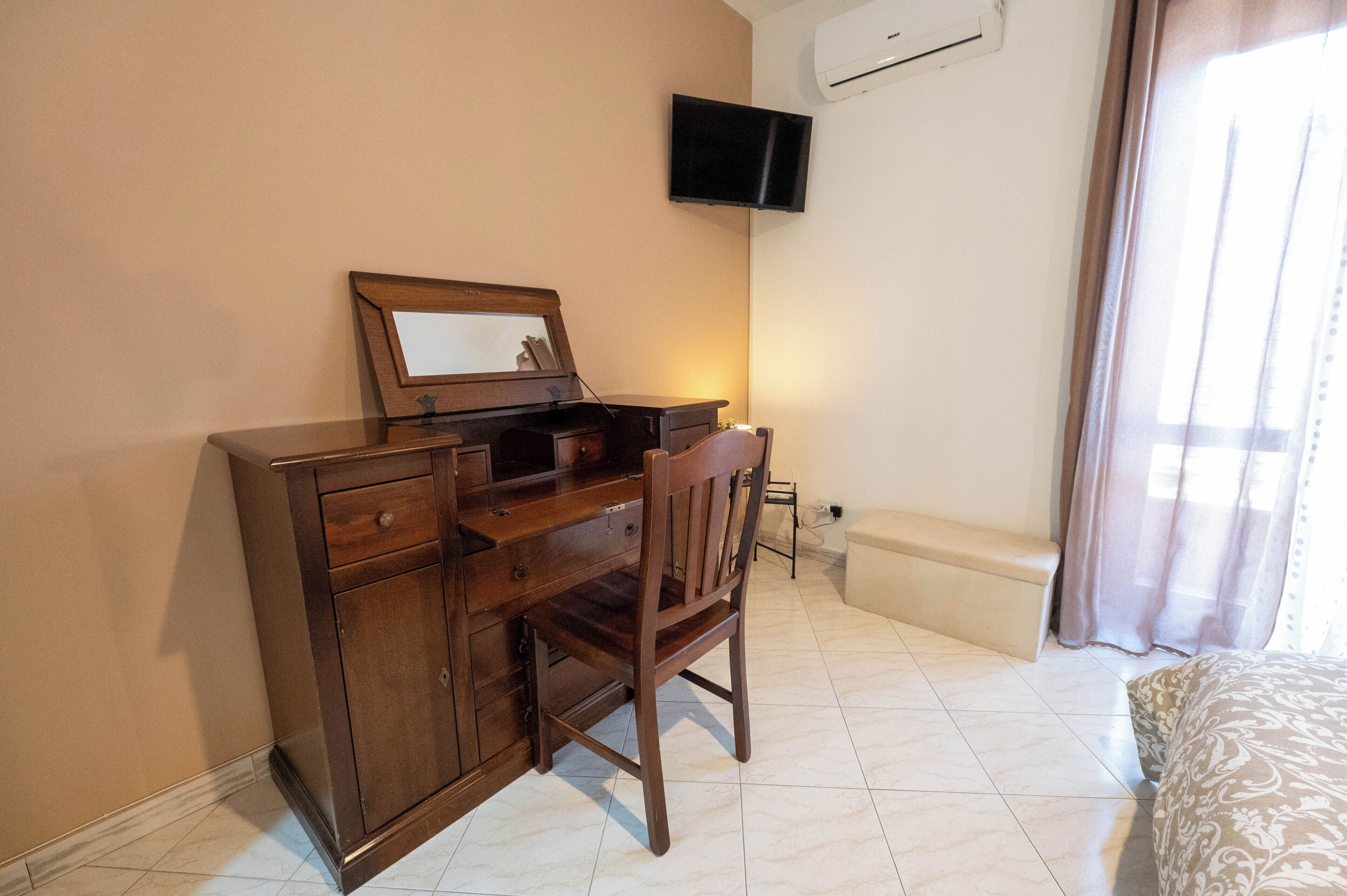 A room with a wooden desk, a chair, a television mounted on the wall, and a window with curtains.