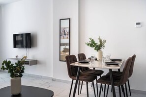 A modern dining area with a table set for four, a flat-screen TV mounted on the wall, and a large mirror reflecting the room. - Opal Signature Studio (Larnaca)