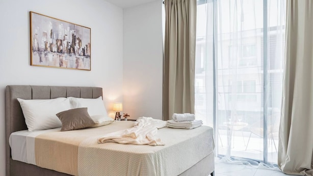 Quarto de hotel com cama, mesa de cabeceira, luminária, janela com cortinas e uma pintura com vista para a cidade.