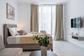 A modern hotel room with a bed, a television, a plant, and a window with curtains. - Opal Signature Studio (Larnaca)