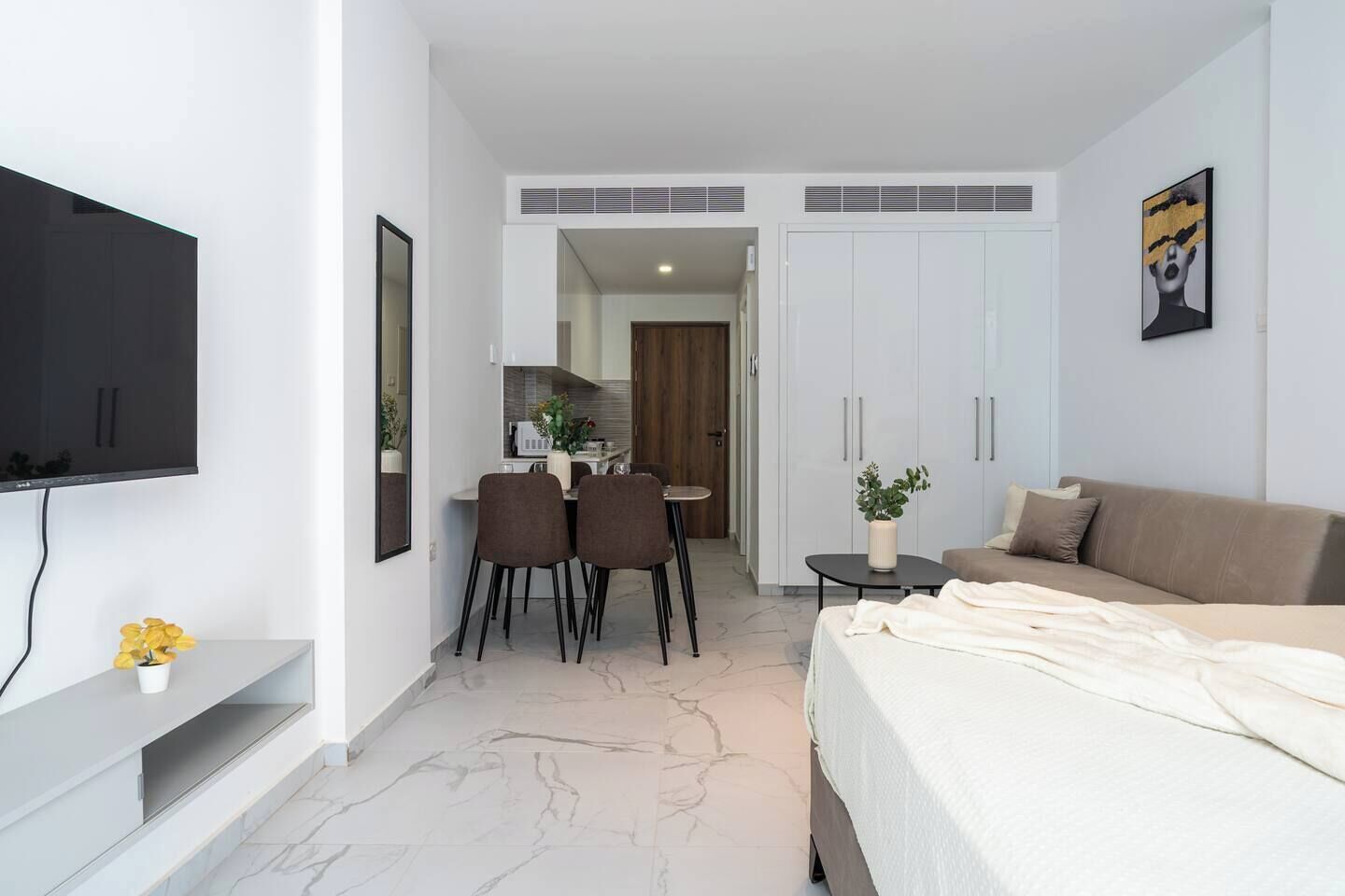 A modern hotel room with a bed, a TV, a dining area, and a kitchenette.