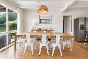 A modern dining area with a wooden table and white chairs, a hanging light fixture, and a view of greenery outside. - Moonbay by Ready Set Stay (Rye)