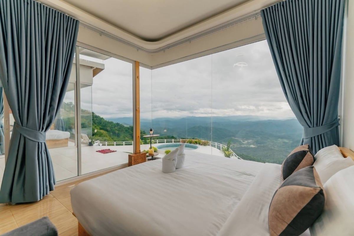 A bedroom with a large bed, a view of a mountainous landscape, and a balcony with a table and chairs.