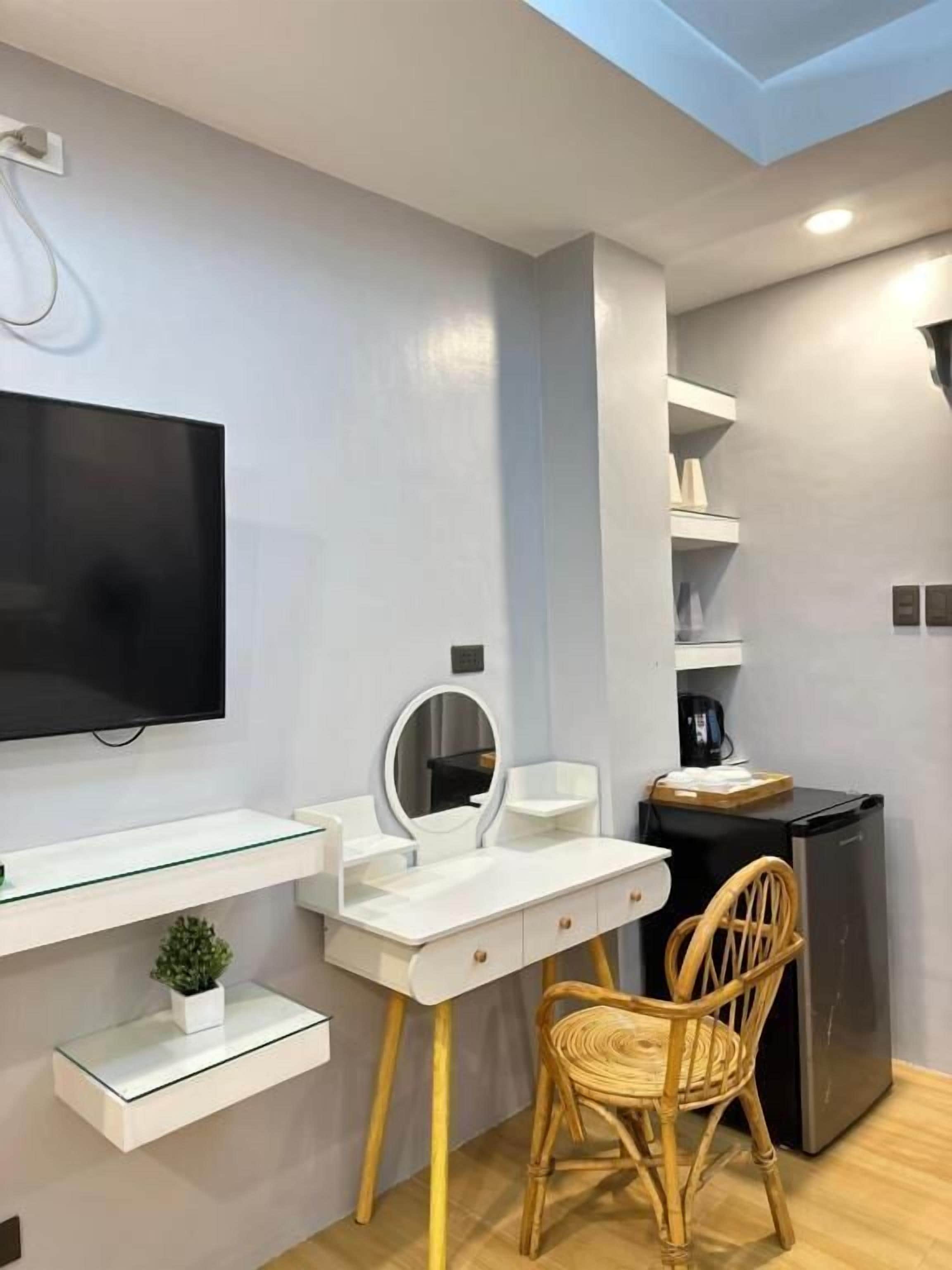 Quarto moderno com televisão de tela plana na parede, toucador branco com espelho, uma pequena planta em vaso, uma cadeira de vime e um frigorífico compacto.