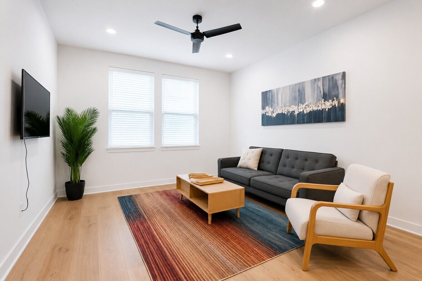 Una sala de estar moderna con un sofá gris, una mesa de centro de madera, una sola silla, un televisor y un ventilador de techo.