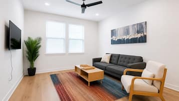 Una sala de estar moderna con un sofá gris, una mesa de centro de madera, una sola silla, un televisor y un ventilador de techo.