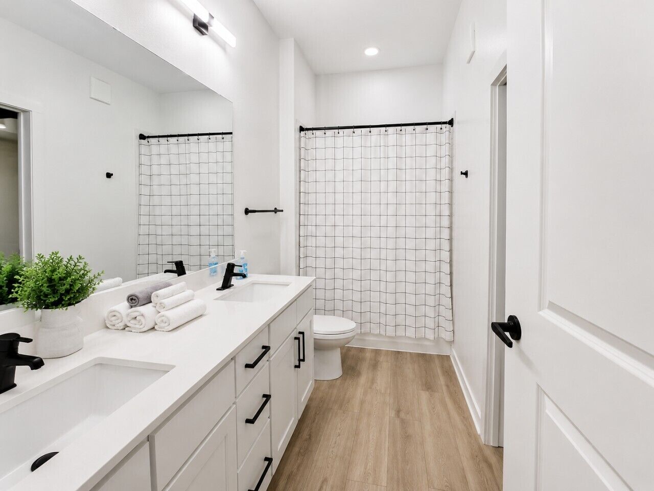Un baño moderno con lavamanos, espejo y retrete blancos, una ducha con cortina de diseño de cuadrícula y suelo de madera.