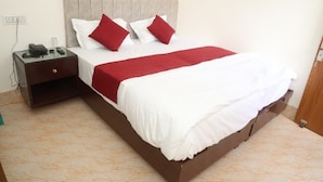 A neatly made bed with white and red bedding, a wooden nightstand with a phone and remote, and a simple wall socket.