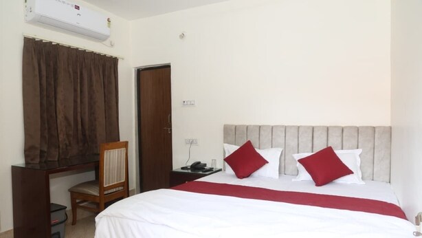 A hotel room with a bed, a desk with a chair, a telephone, and an air conditioning unit.