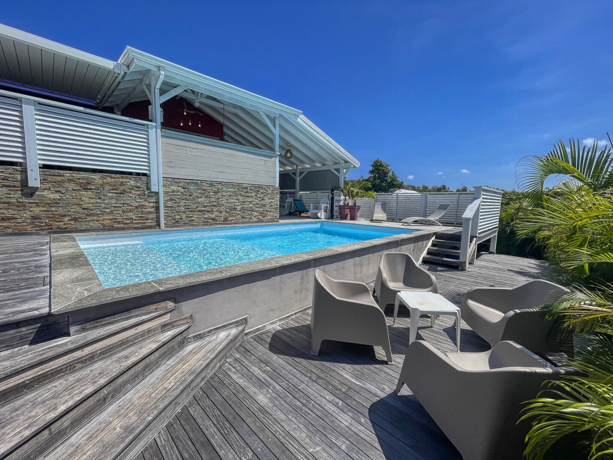 A swimming pool with a wooden deck, lounge chairs, and a covered patio area.