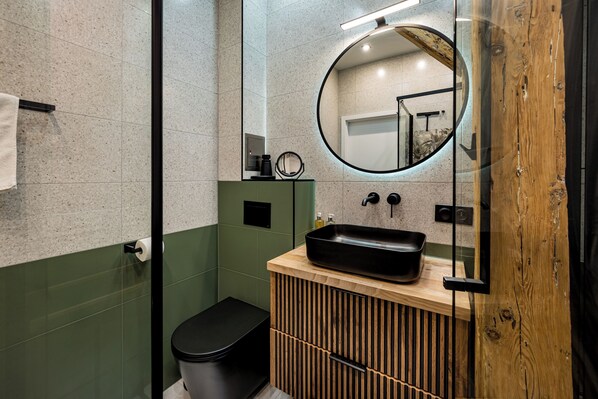 Un baño moderno con cabina de ducha de vidrio, inodoro negro, tocador de madera con lavamanos negro y espejo con marco de madera.