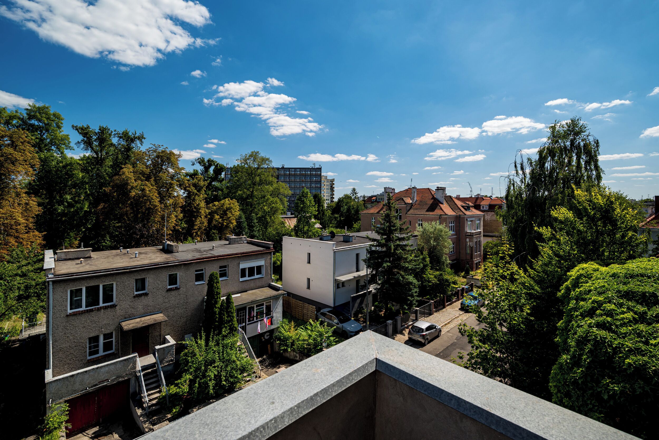 A residential area with various buildings, including a modern apartment and a traditional house with a red roof, surrounded by trees and under a clear sky.
