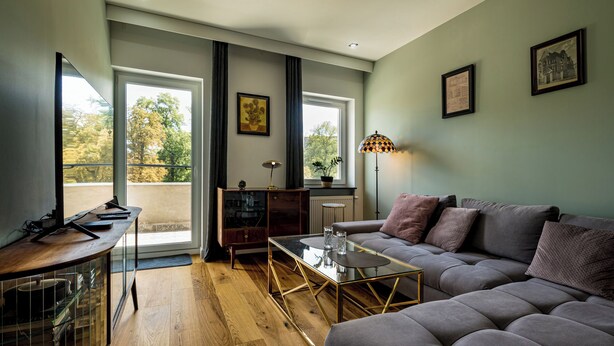 Una sala de estar moderna con un sofá, una mesa de centro y un escritorio con una computadora. Hay ventanas con cortinas, una lámpara y cuadros en la pared.