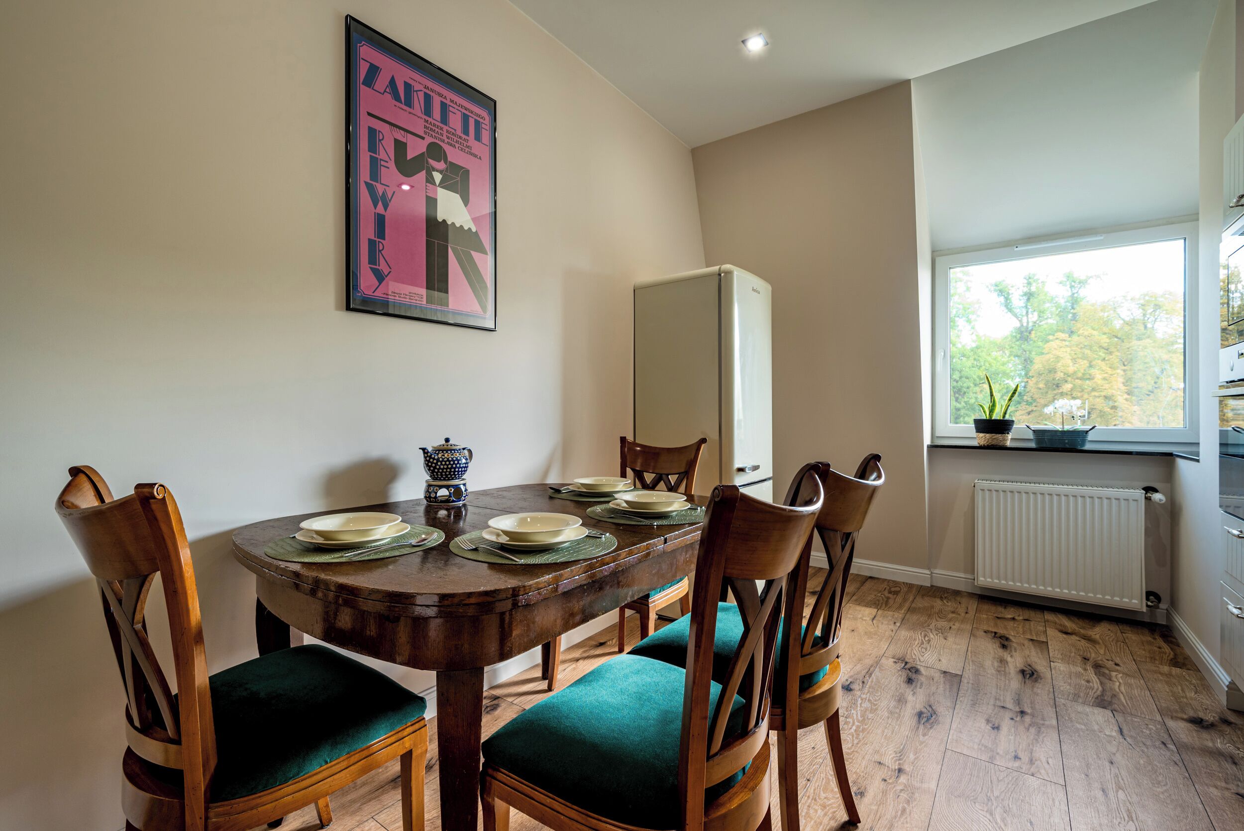 A dining area with a wooden table set for four, surrounded by chairs with green cushions. A poster is mounted on the wall above the table.