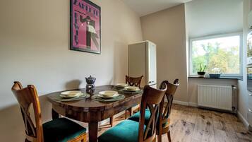 A dining area with a wooden table set for four, surrounded by chairs with green cushions. A poster is mounted on the wall above the table.