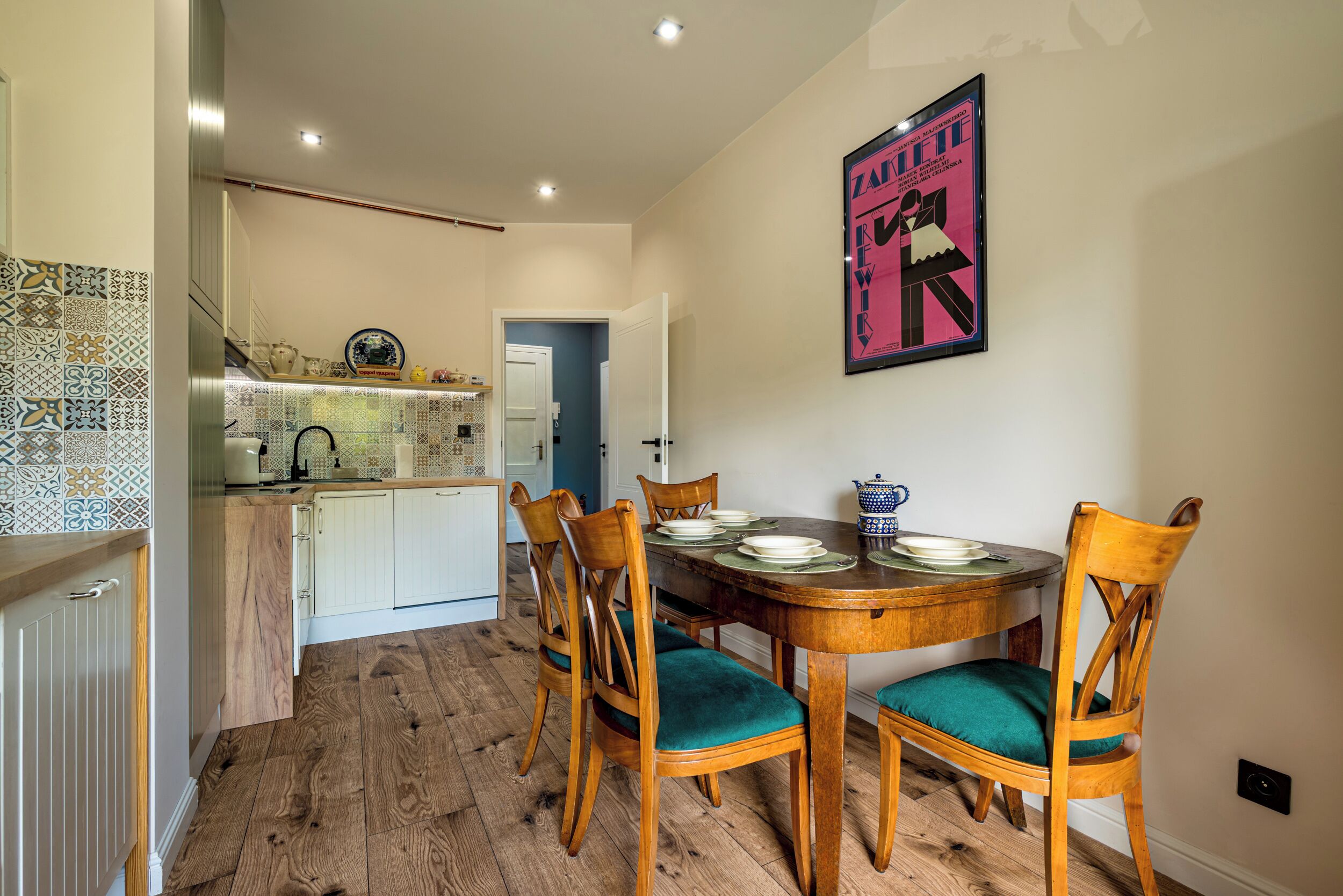A dining area with a wooden table and chairs, a kitchen in the background, and a poster on the wall.