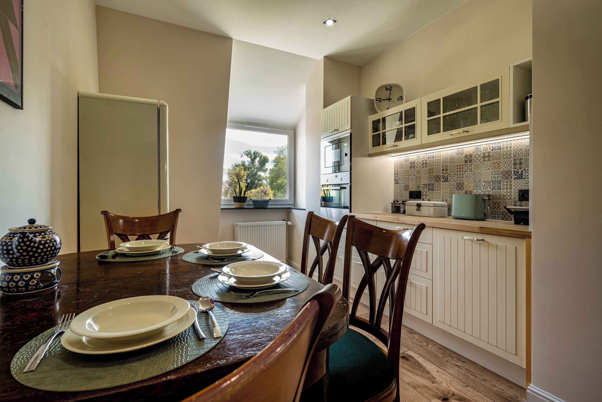 A dining area with a wooden table set for four, a kitchen with white cabinets and patterned backsplash, and a window providing a view of trees.