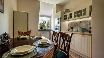 A dining area with a wooden table set for four, a kitchen with white cabinets and patterned backsplash, and a window providing a view of trees.