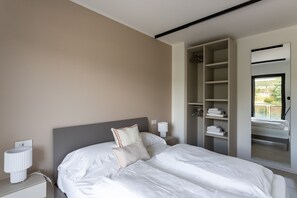 Ein Hotelzimmer mit einem Bett, einem Nachttisch, einer Lampe und einem Kleiderschrank mit darin hängender Kleidung.