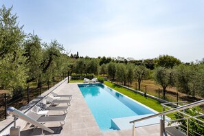 Ein rechteckiger Swimmingpool mit glasklar blauem Wasser, umgeben von einer niedrigen Steinmauer. Weiße Liegestühle sind entlang des Beckenrandes aufgereiht, und eine Metalltreppe mit Geländer führt hinab zum Pool. Das Gebiet wird von einem schwarzen Metallzaun begrenzt, und dahinter erstreckt sich vielfältiges Grün mit Bäumen und Sträuchern.