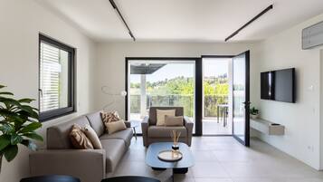 A modern living room with a sofa, armchairs, a coffee table, and a TV. There is a balcony with a view of greenery.