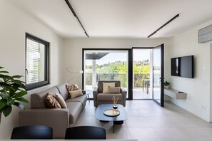 Ein modernes Wohnzimmer mit einer Couch, Sesseln, einem Couchtisch und einem Fernseher. Ein Balkon bietet Blick auf viel Grün.