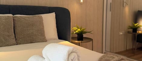 Una cama bien hecha con toallas enrolladas y un edredón mullido, una mesita con una planta, y una puerta corrediza que da a otra habitación.
