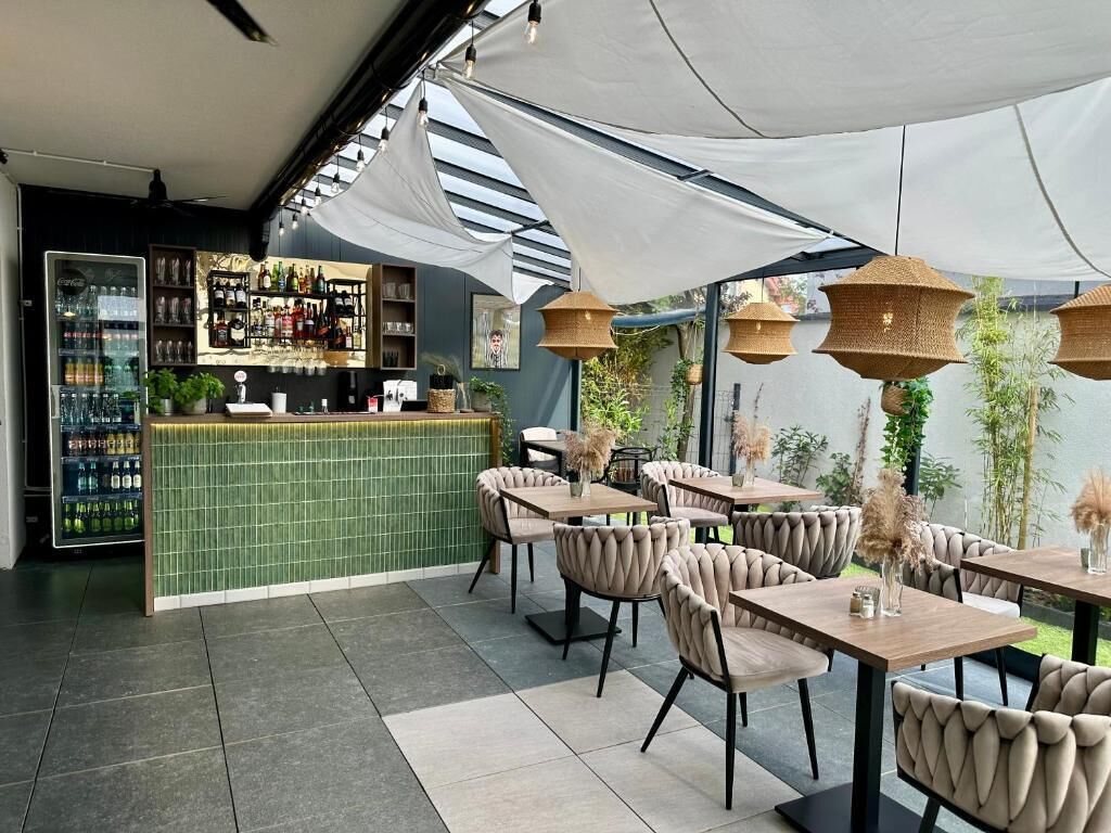 An outdoor seating area with tables and chairs, a bar with shelves of bottles, and a green tiled counter.