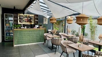 An outdoor seating area with tables and chairs, a bar with shelves of bottles, and a green tiled counter.