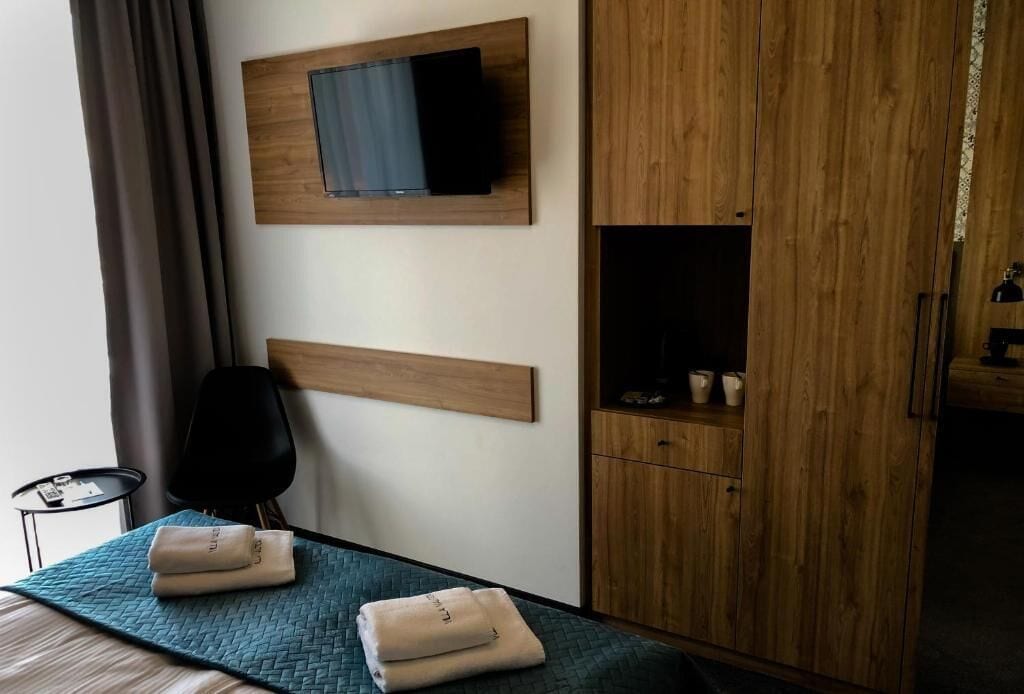 A hotel room with a bed, a TV mounted on the wall, a wooden wardrobe, and a small table with a chair.