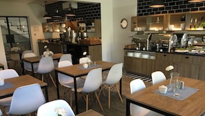 Un café moderno con mesas de madera y sillas blancas, una máquina de café y una zona de bufé con alimentos.