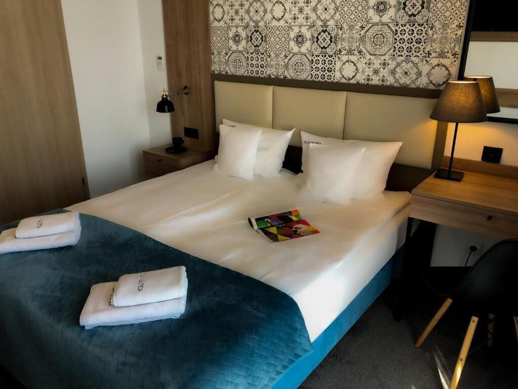 Una cama bien hecha con ropa blanca y un detalle en azul, una mesita de noche con una lámpara, una pared con papel tapiz estampado y un escritorio pequeño con una silla.