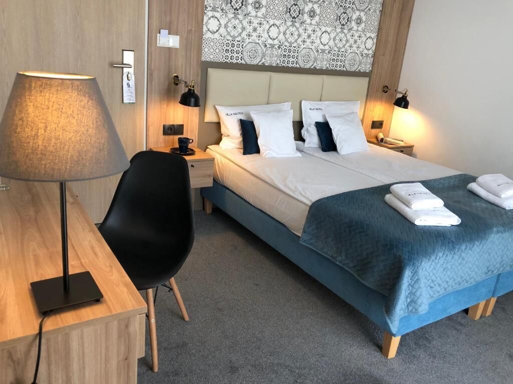A hotel room with a bed, bedside lamps, a desk with a lamp, a chair, and a wall tapestry.