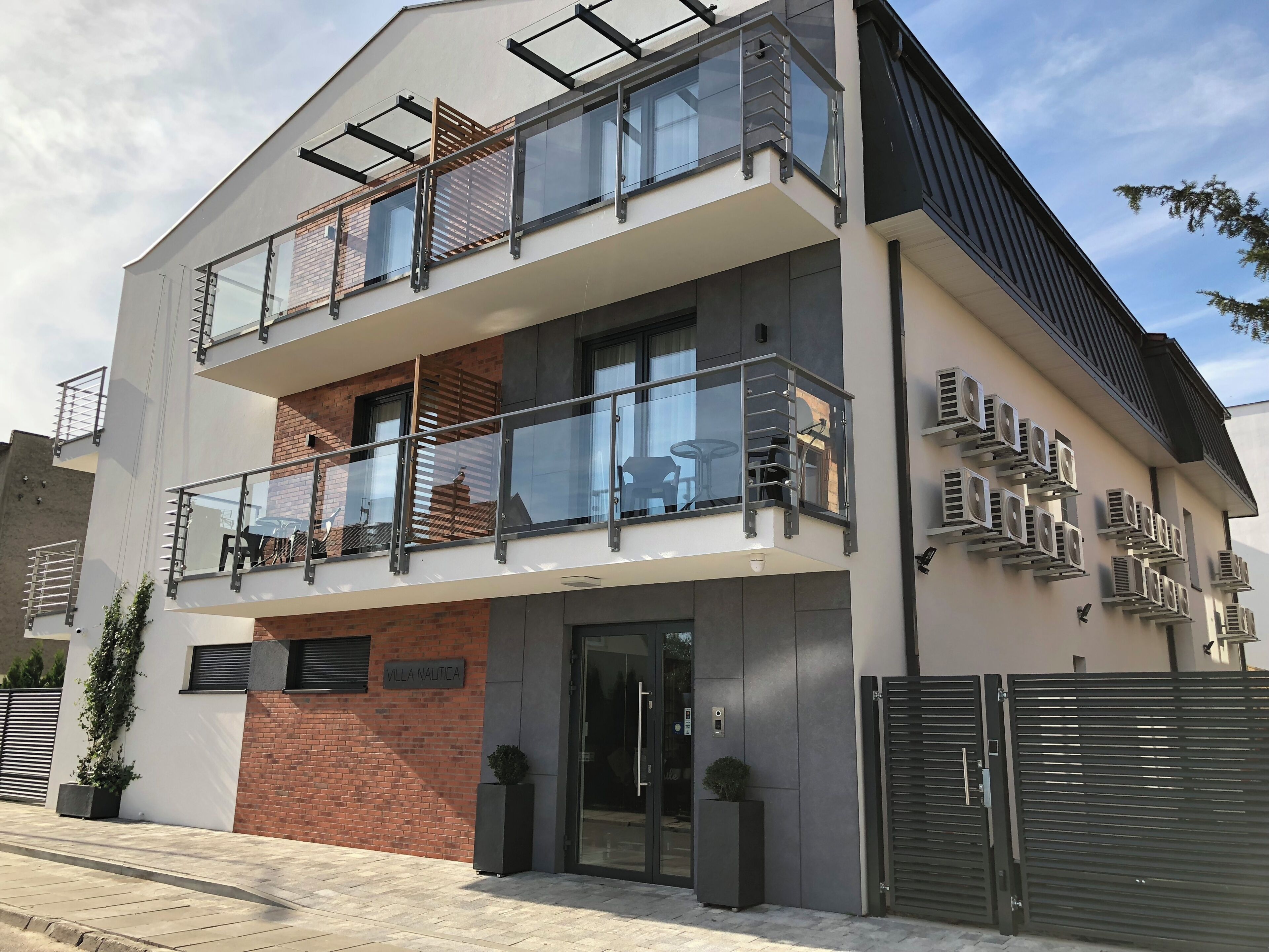 Ein modernes, zweistöckiges Gebäude mit einem Glasbalkon, einer Schilder an der Fassade mit der Aufschrift 'Villa Nautica' und mehreren Klimaanlagen an der Außenwand.