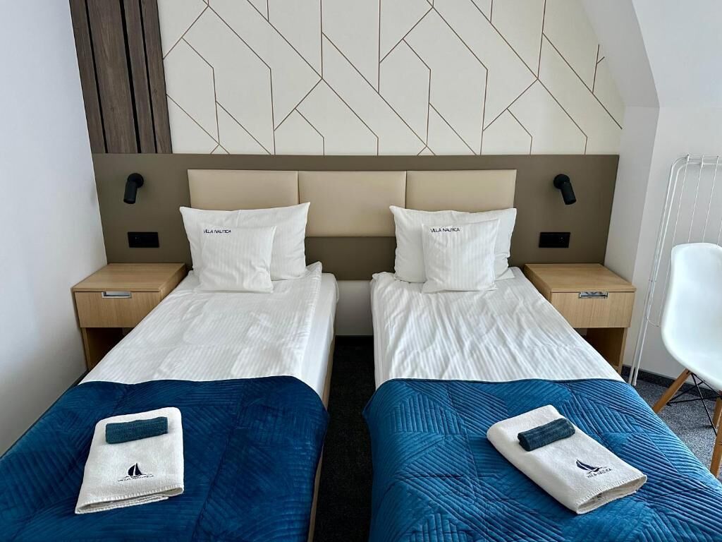 A hotel room with two beds, each with a pillow labeled 'VILLA NAUTICA,' a wooden headboard, and a wall with a geometric pattern.