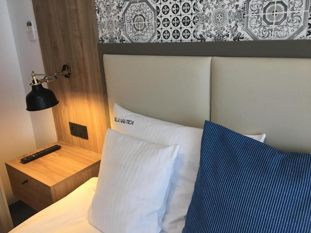 A hotel room with a bed, a nightstand, a remote control, and a wall-mounted lamp.