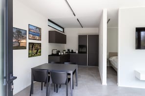 A modern kitchen with dark cabinetry, a dining area with a table and chairs, and a bed in the adjacent room.