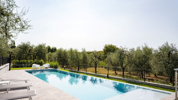 A rectangular swimming pool with clear blue water, surrounded by a low stone wall. There are white lounge chairs and a small table on the poolside.