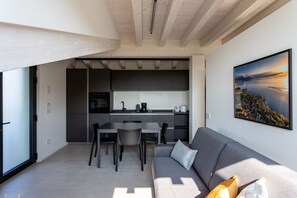 A modern kitchen with dark cabinets, a dining area with a gray sofa and chairs, and a large window. - Casa Valtenesi - 3 Sensi (Puegnago sul Garda)