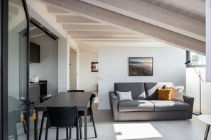 Een moderne woonkamer met een grijze bank, zwarte stoelen, een eettafel en een keuken met een zwart kastje en witte apparaten.